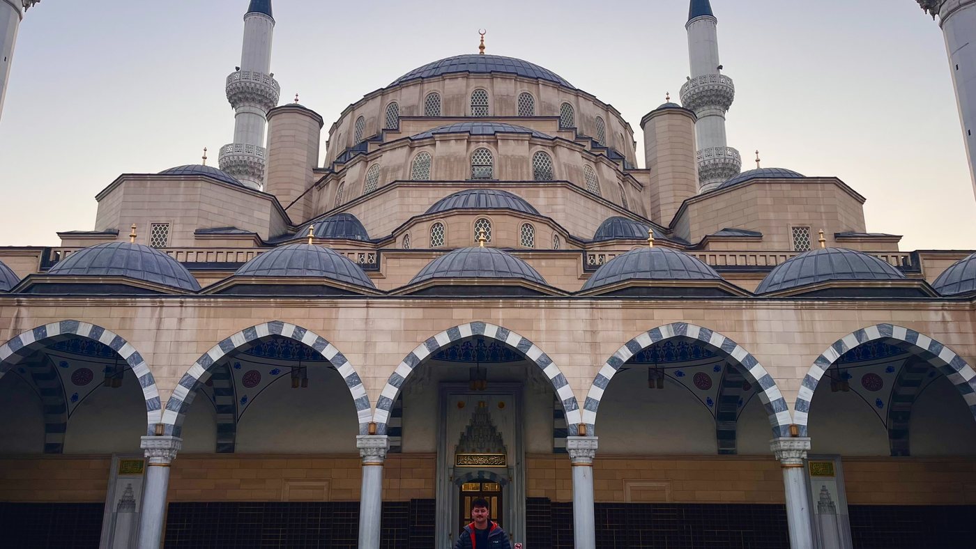 The Central Mosque in Bishkek at dawn — domes, twin minarets and a colonnade of arches, with a visitor standing in the empty courtyard for scale.