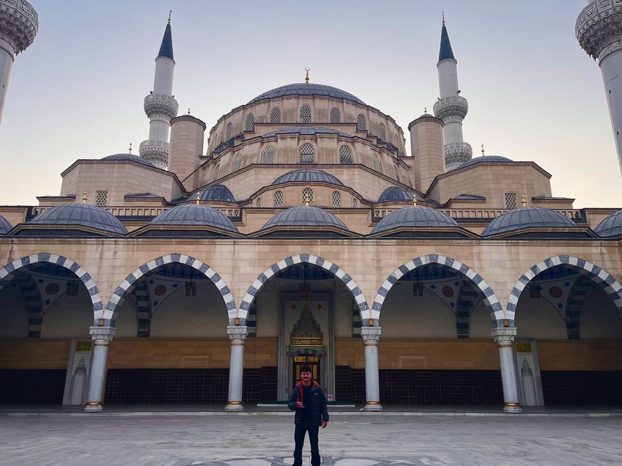 The Central Mosque in Bishkek at dawn — domes, twin minarets and a colonnade of arches, with a visitor standing in the empty courtyard for scale.
