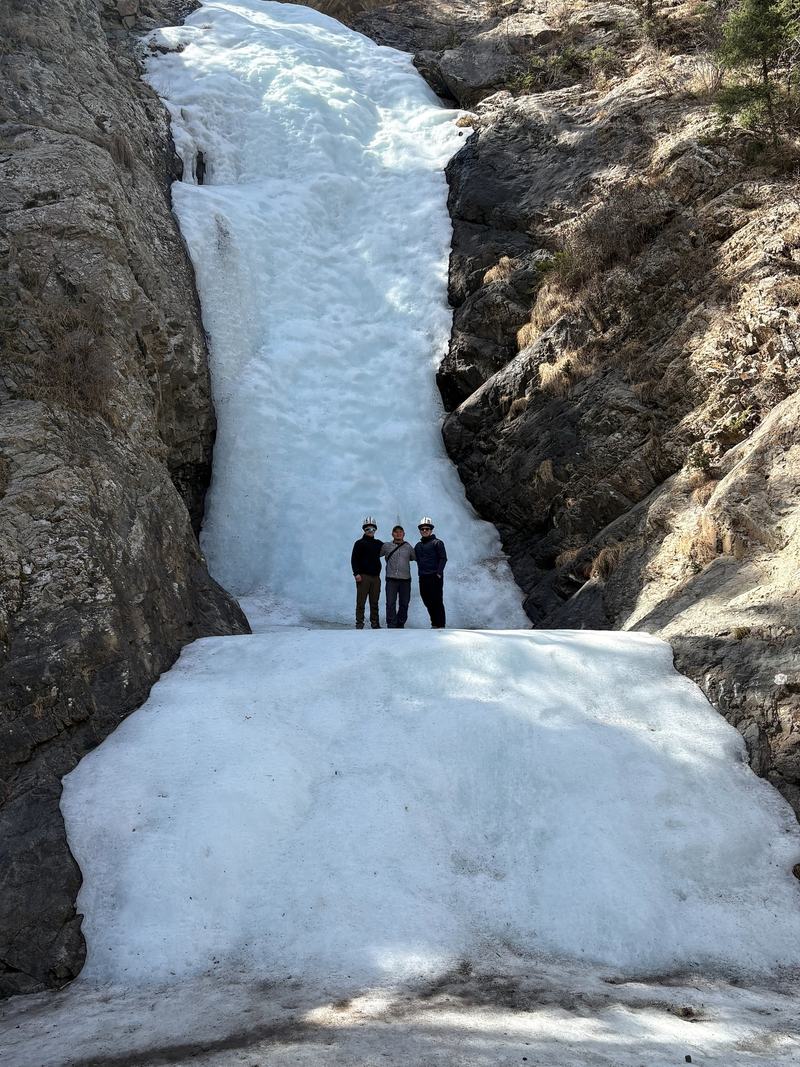 Three men in white Kyrgyz kalpak hats standing at the foot of a frozen waterfall filling a narrow rock gorge.