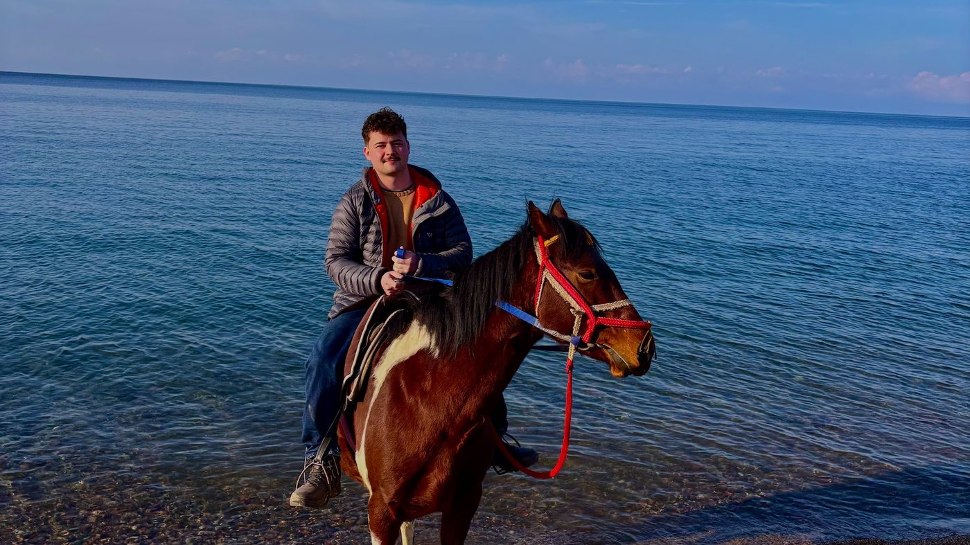 A traveller riding a horse along the pebble shore of Issyk-Kul, the horse standing in the shallows with the open lake and a clear horizon behind.
