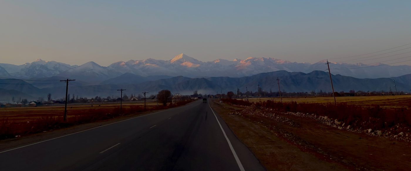 An empty two-lane road running straight towards the snow-covered peaks of the Tian Shan at dusk, with autumn fields and telegraph poles on either side.