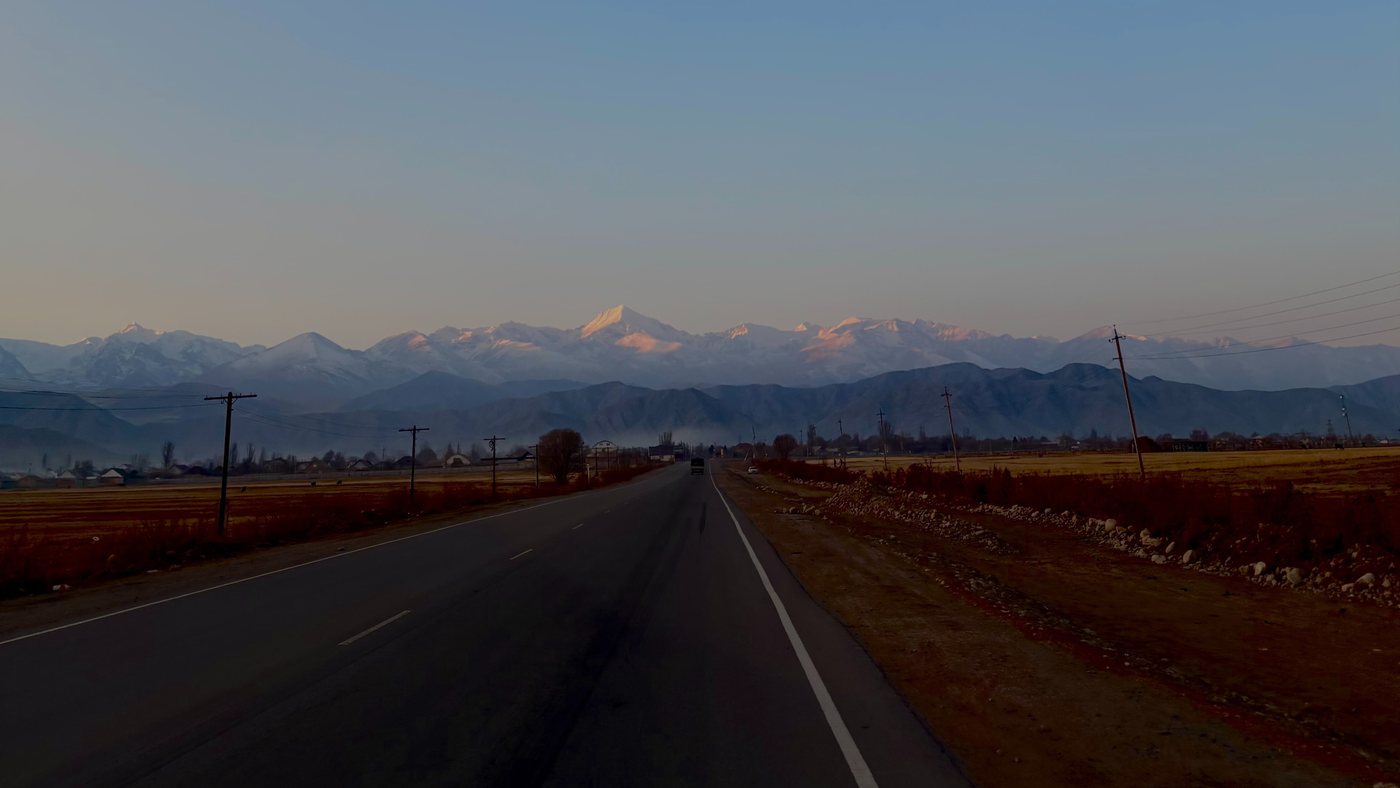 An empty two-lane road running straight towards the snow-covered peaks of the Tian Shan at dusk, with autumn fields and telegraph poles on either side.