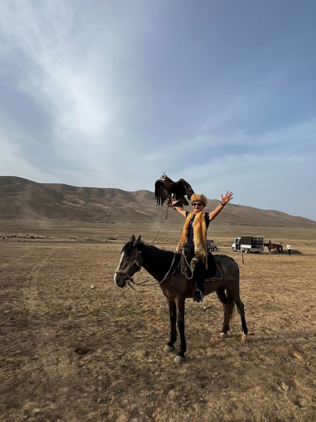 A traveller on horseback in a Kyrgyz kalpak hat holding a golden eagle on a gloved arm, one hand raised, on open ground below dry hills.